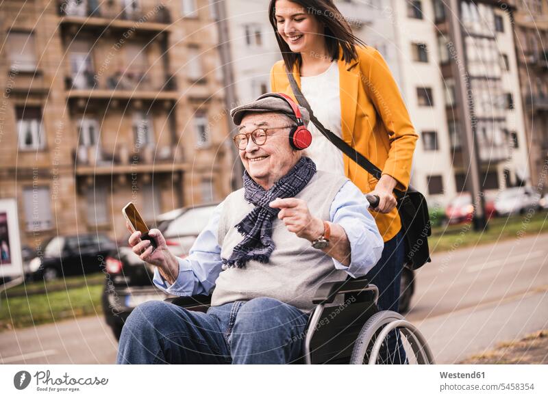 Laughing young woman pushing happy senior man with headphones and smartphone in wheelchair human human being human beings humans person persons
