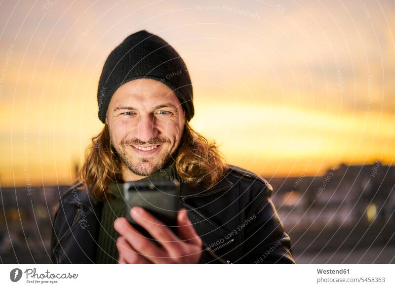 Portrait of bearded young man wearing wooly hat looking at mobile phone by sunset Smartphone iPhone Smartphones portrait portraits eyeing woolly hat Wooly Hat