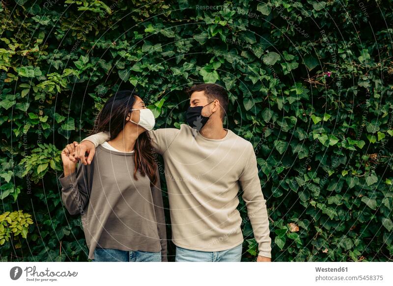 Young couple with arm around standing against leaves in park during pandemic color image colour image outdoors location shots outdoor shot outdoor shots day