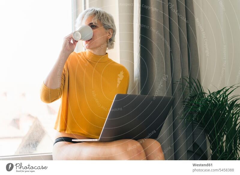 Woman sitting at the window at home with laptop drinking coffee from cup human human being human beings humans person persons celibate celibates singles