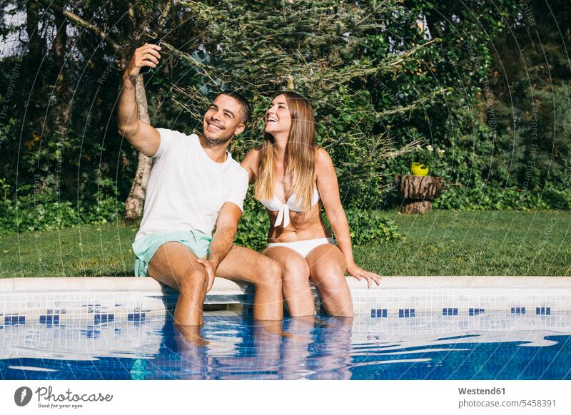 Smiling man taking selfie with girlfriend over smart phone while sitting against trees at poolside color image colour image Spain day daylight shot