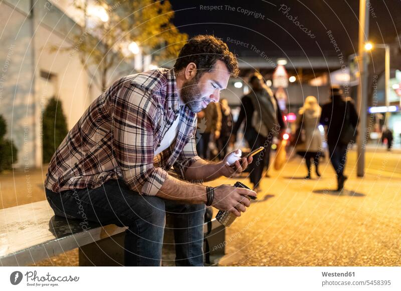 UK, London, smiling man sitting on a bench and looking at his phone by night men males smile benches mobile phone mobiles mobile phones Cellphone cell phone