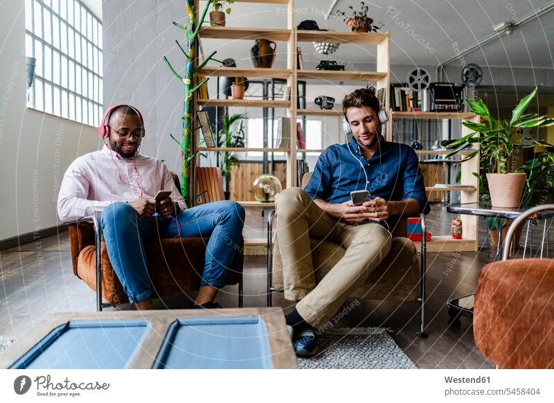 Two young men with cell phones and headphones sitting in armchairs in a loft Seated man males mobile phone mobiles mobile phones Cellphone headset lofts