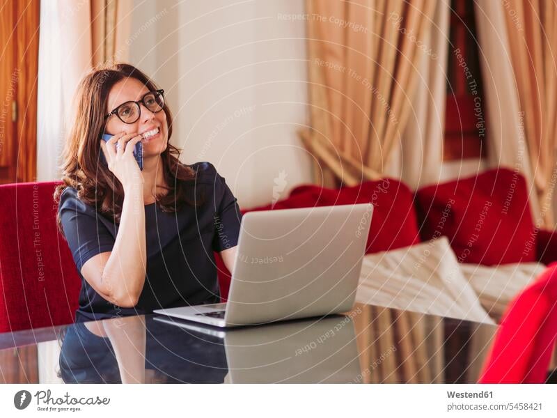 Smiling businesswoman talking on mobile phone while sitting with laptop at table in living room color image colour image Spain indoors indoor shot indoor shots