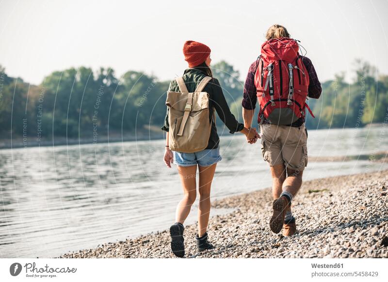 Rear view of young couple with backpacks walking hand in hand at the riverside rucksacks back-packs going River Rivers riverbank twosomes partnership couples