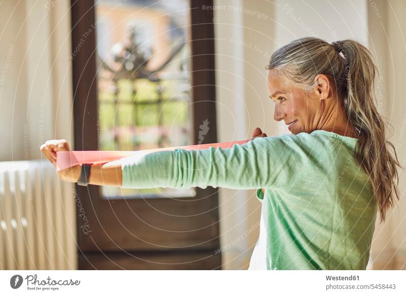 Smiling woman exercising with resistance band in living room Germany indoors indoor shot indoor shots interior interior view Interiors day daylight shot
