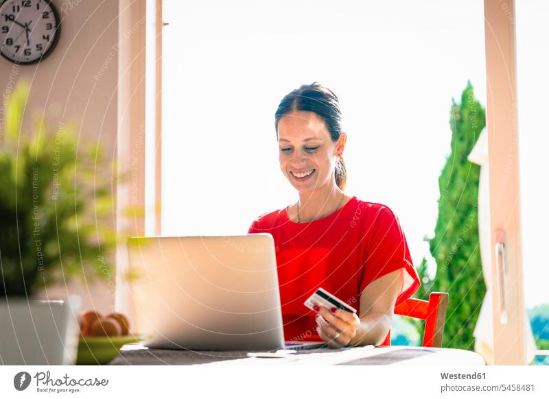 Smiling beautiful woman holding credit card while using laptop at dining table against window color image colour image indoors indoor shot indoor shots interior