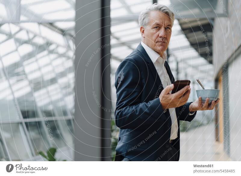 Senior businessman holding smartphone and muesli bowl in office human human being human beings humans person persons caucasian appearance caucasian ethnicity