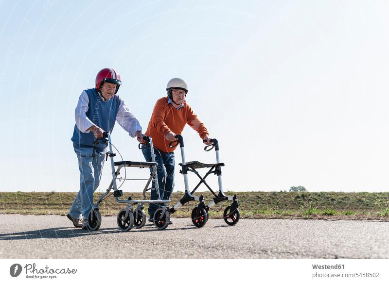 Two old friends wearing safety helmets, competing in a wheeled walker race Wheeled Walker running competition country road rural road rural roads country roads
