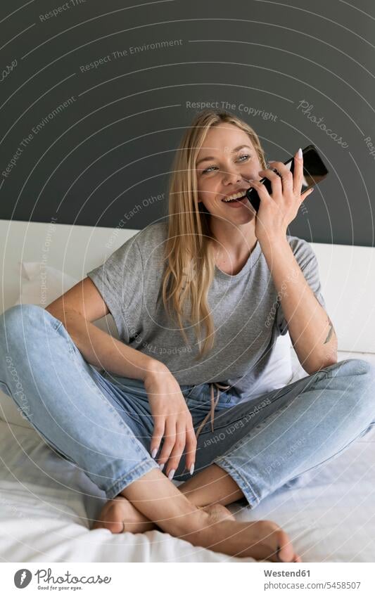 Happy young woman sitting on bed using cell phone Seated happiness happy females women mobile phone mobiles mobile phones Cellphone cell phones beds Adults