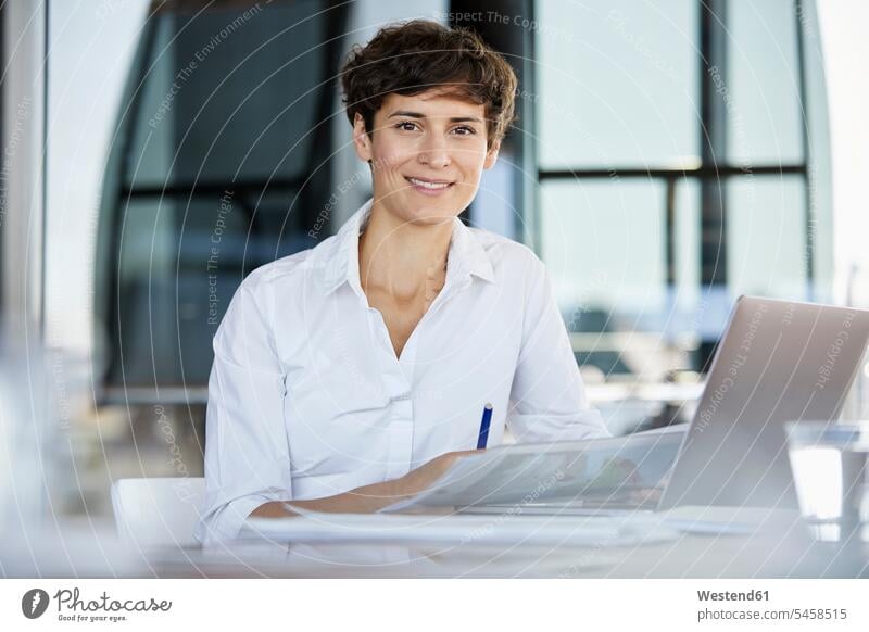 Portrait of confident businesswoman sitting at desk in office with laptop and document desks offices office room office rooms Seated Laptop Computers laptops