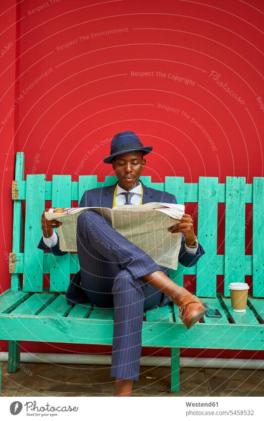 Young businessman wearing old-fashioned suit and hat sitting on a green bench reading the newspaper human human being human beings humans person persons African