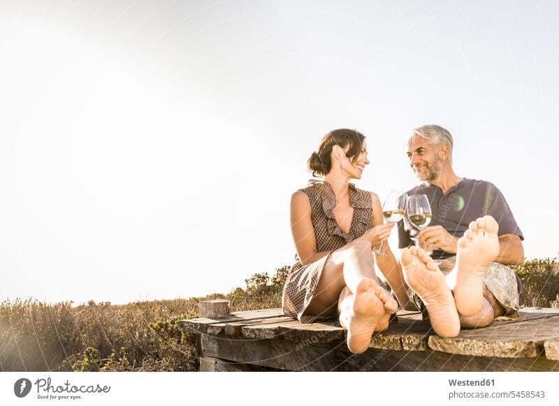 Happy couple sitting on boardwalk at the coast enjoying a glass of white wine touristic tourists smile Seated drink in the evening Late Evening seasons