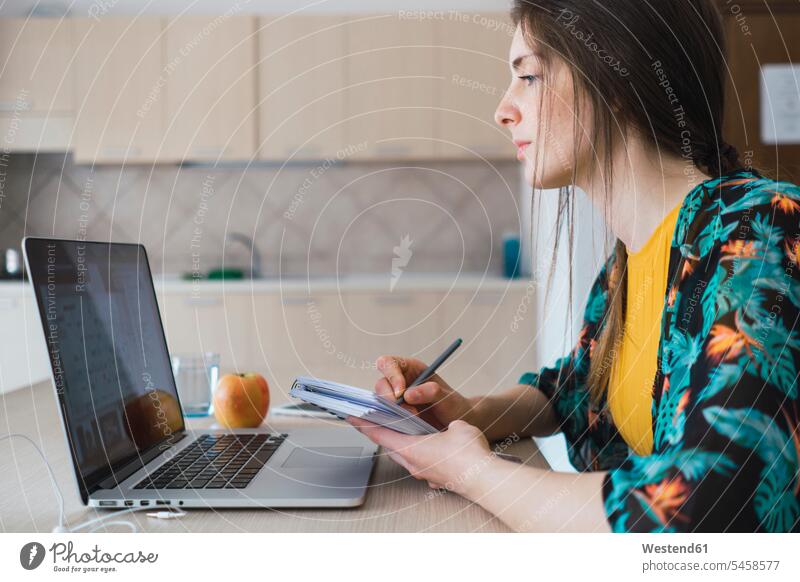 Young woman sitting at table at home using laptop and taking notes Laptop Computers laptops notebook Table Tables making a note note taking females women