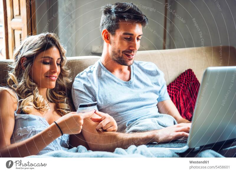 Young couple sitting on couch at home with credit card and laptop finance financial credit cards debit card direct payment couches settee settees sofa sofas