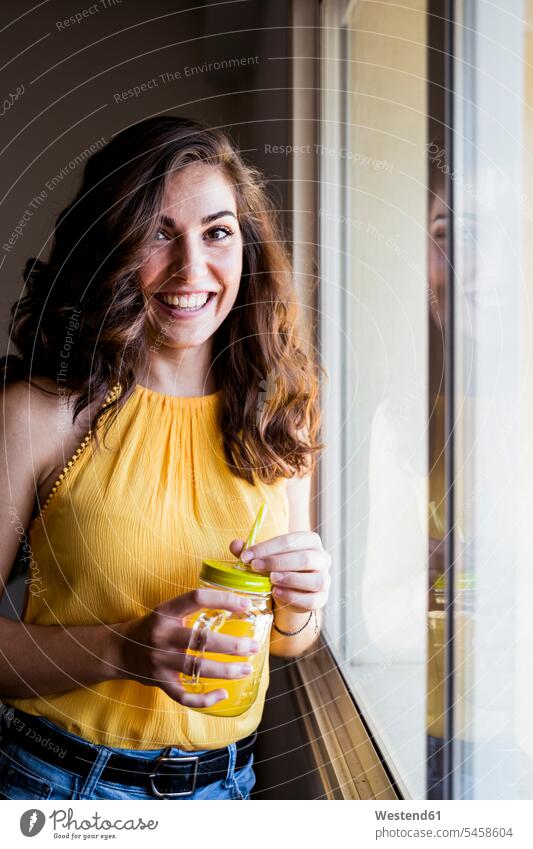 Beautiful young woman holding mason jar with juice while smiling by window at home color image colour image Spain leisure activity leisure activities free time
