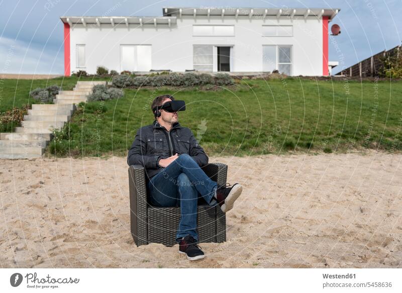 Man sitting in armchair on the beach wearing VR glasses Arm Chairs armchairs virtual reality man men males beaches specs Eye Glasses spectacles Eyeglasses