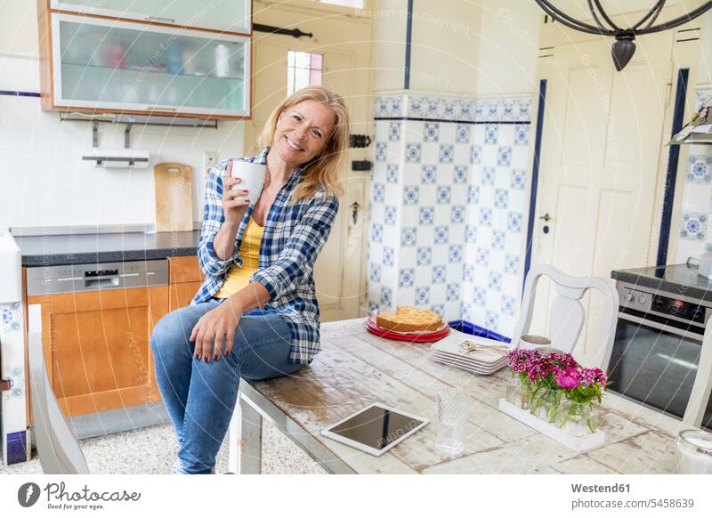 Portrait of smiling mature woman with coffee mug sitting on dining table in the kitchen human human being human beings humans person persons