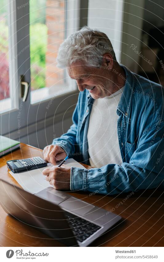 Happy senior man writing on paper while using calculator and laptop at home color image colour image indoors indoor shot indoor shots interior interior view