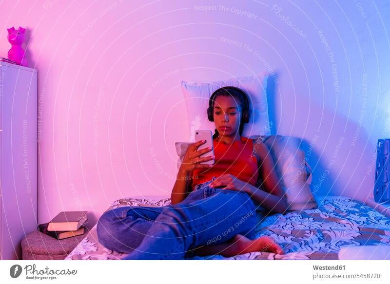 Teenage girl lying on bed using smart phone to listen music through headphone at home color image colour image indoors indoor shot indoor shots interior