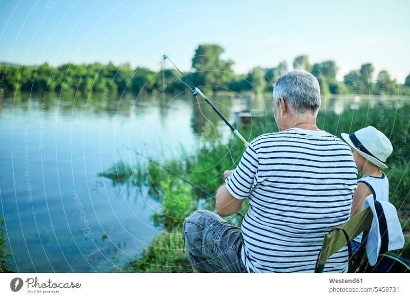 Back view of grandfather and grandson fishing together at lakeshore Lakeshore Lake Shore lakeside grandsons grandpas granddads grandfathers angling water's edge