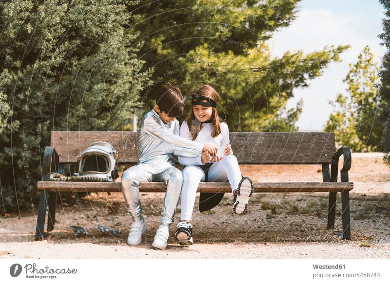 Two kids in astronaut and superhero costumes with mobile phone on park bench friends mate human human being human beings humans person persons braver bravers