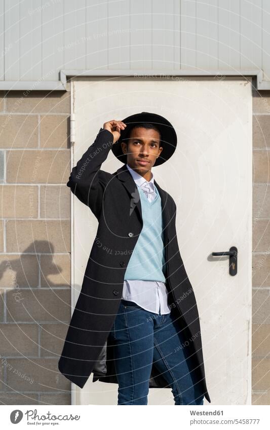 Fashionable young man holding hat while standing against door on sunny day color image colour image outdoors location shots outdoor shot outdoor shots