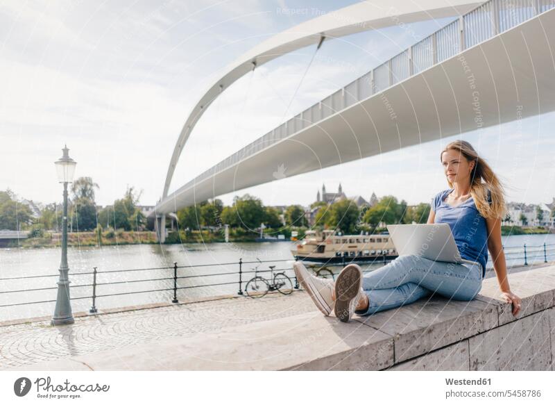 Netherlands, Maastricht, young woman sitting on a wall at the riverside with laptop Laptop Computers laptops notebook walls Seated riverbank females women River