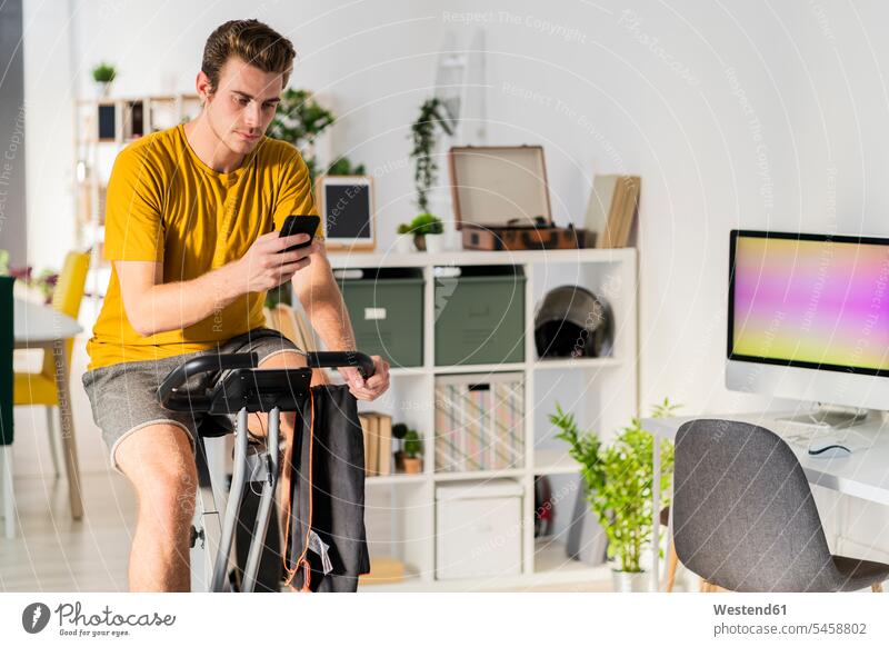 Man using mobile phone while cycling on exercise equipment at home color image colour image indoors indoor shot indoor shots interior interior view Interiors