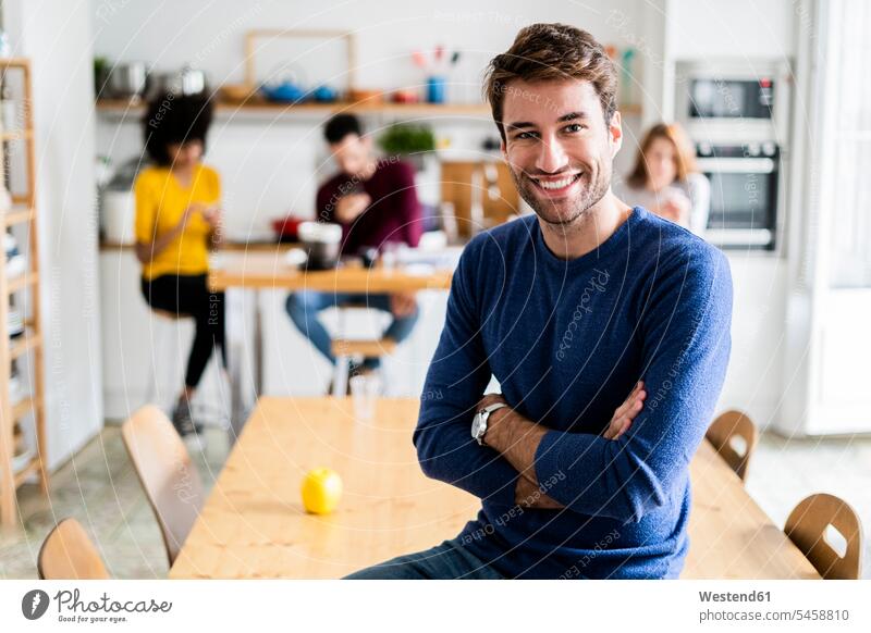 Portrait of smiling man at dining table at home with friends in background mate students jumper sweater Sweaters Tables Dining Tables Dinner Table wood