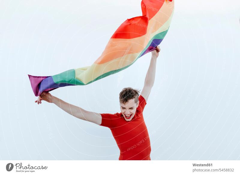 Non-binary person waving multi colored flag while standing against white wall color image colour image Non-binary Person Non-binary People non-binary nonbinary