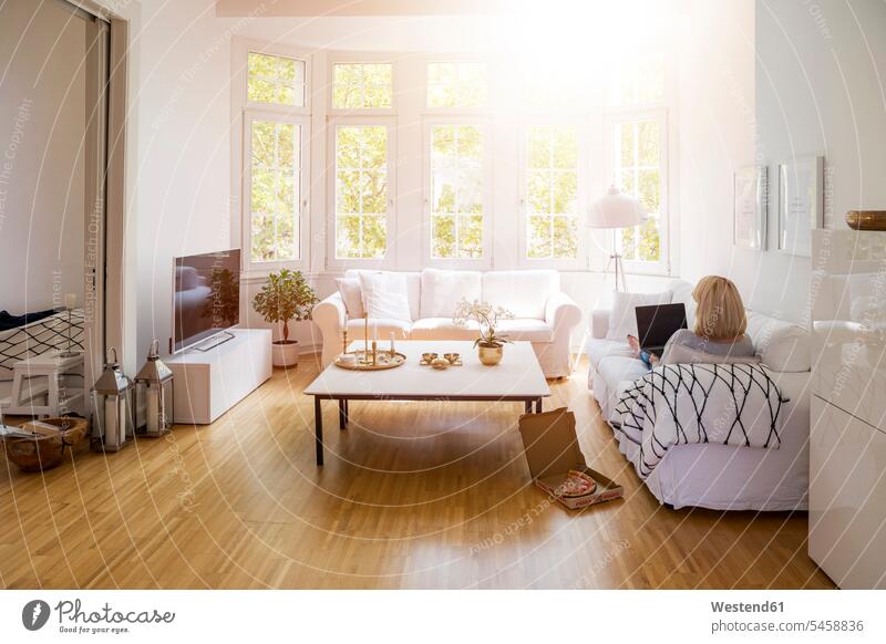 Back view of blond mature woman sitting on couch in the living room using laptop living rooms livingroom settee sofa sofas couches settees blond hair