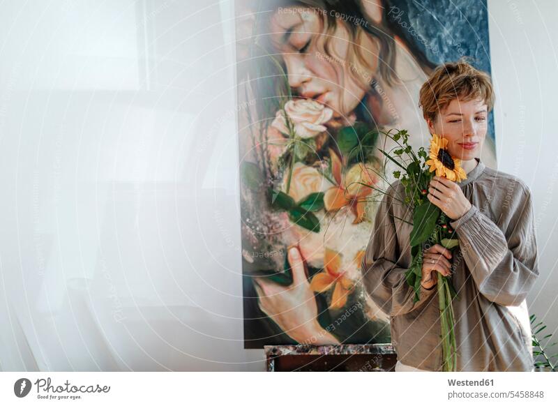 Thoughtful mid adult female artist holding sunflower while standing against artwork in studio color image colour image painter painters indoors indoor shot