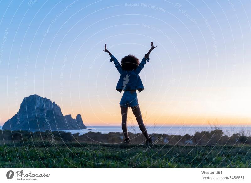Rear view of young woman standing on a viewpoint with raised arms at sunset, Ibiza in the evening Late Evening delight enjoyment Pleasant pleasure indulgence