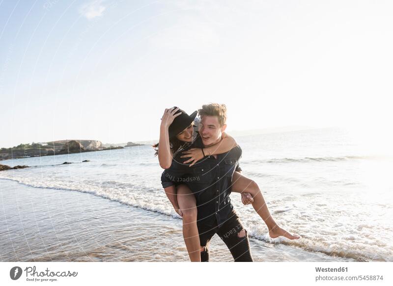 France, Brittany, happy young man carrying girlfriend piggyback at the beach piggy-back pickaback Piggybacking Piggy Back couple twosomes partnership couples
