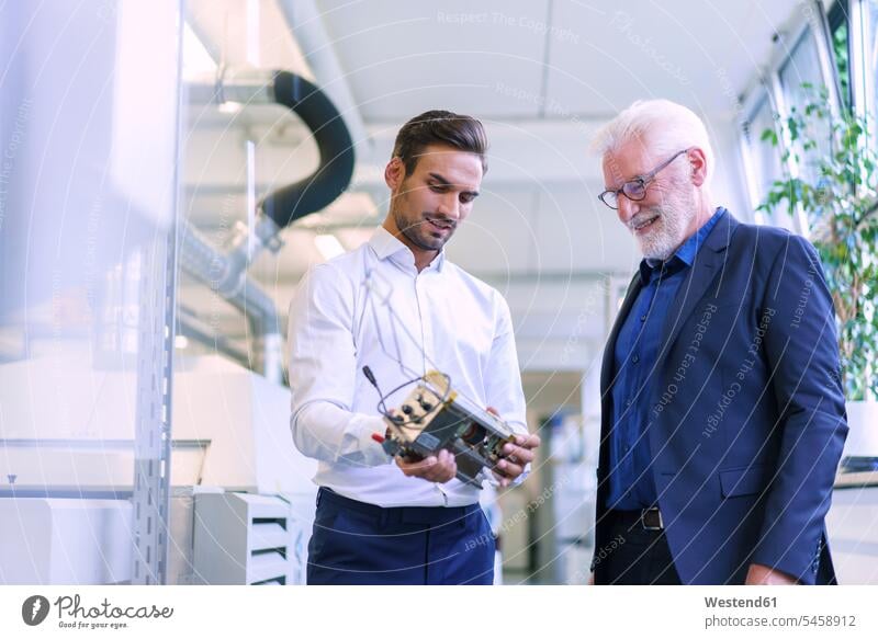 Young male engineer holding machine part while discussing with smiling senior manager at factory color image colour image indoors indoor shot indoor shots