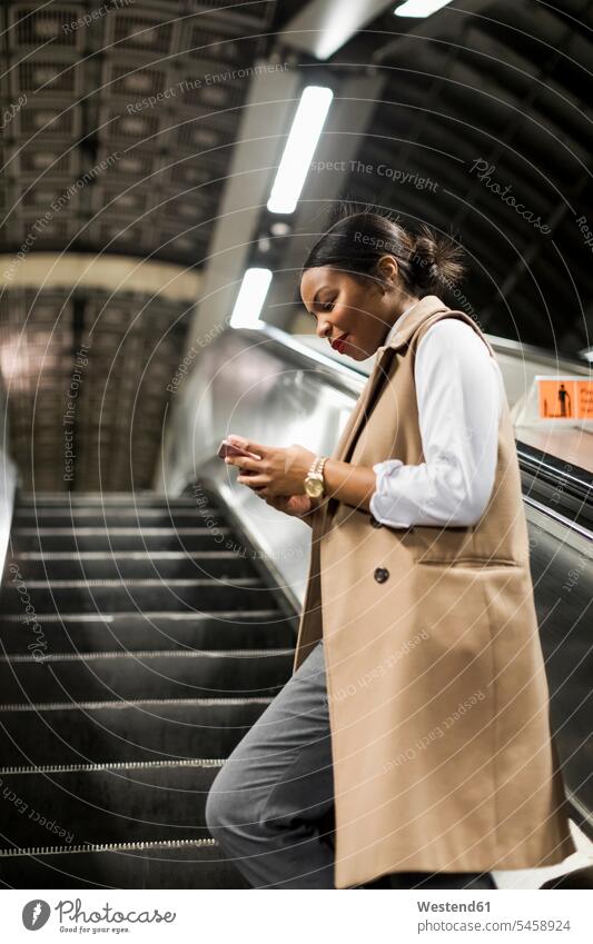 UK, London, smiling businesswoman standing on escalator of underground station looking at cell phone businesswomen business woman business women Smartphone