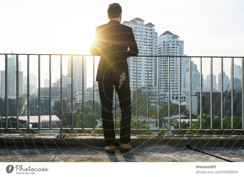 Business man in suit leaning on handrail in sunset on city rooftop Railing Railings Businessman Businessmen Business men standing house top Roof Top roofs