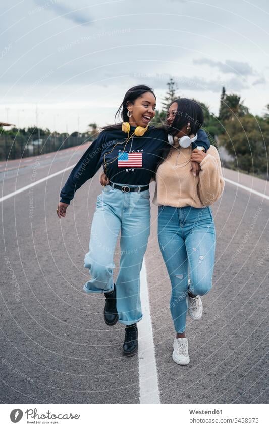 Two happy young women walking on a road friends mate female friend pants Trouser Denim Jeans headphone headset go going smile speak speaking talk embrace