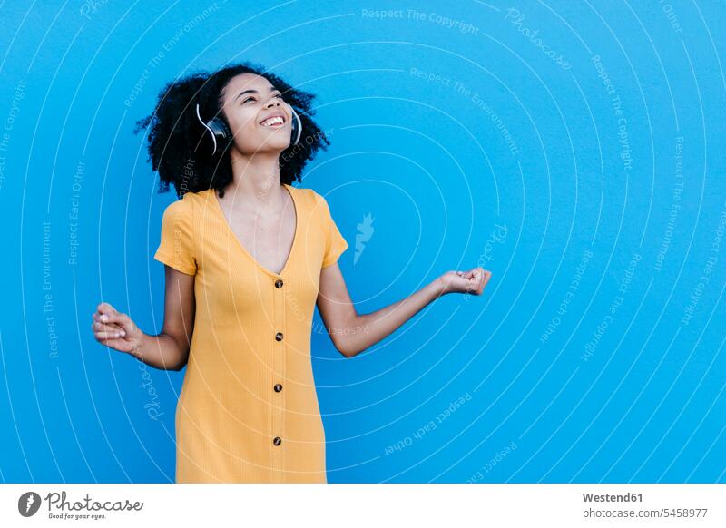 Happy young afro woman enjoying while listening music through headphones against blue wall color image colour image outdoors location shots outdoor shot