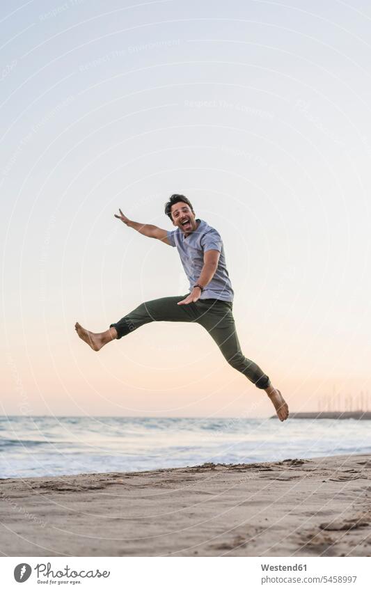 Happy young man jumping in the air on the beach at sunset sunsets sundown beaches men males Leaping jump in the air atmosphere atmospheric mood moody