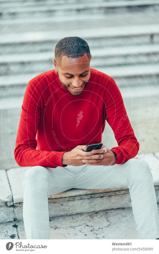 Smilig young man wearing red pullover sitting on stairs using cell phone stairway sweater jumper Sweaters use Smartphone iPhone Smartphones smiling smile men