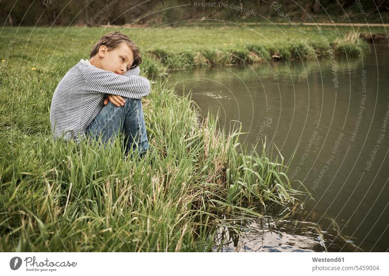 Side view of lonely boy sitting at riverbank while thinking in forest color image colour image Germany outdoors location shots outdoor shot outdoor shots day