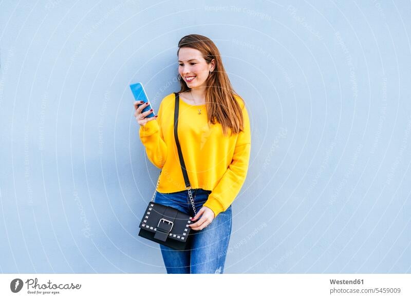Smiling young woman standing at a wall using cell phone mobile phone mobiles mobile phones Cellphone cell phones smiling smile females women walls telephones