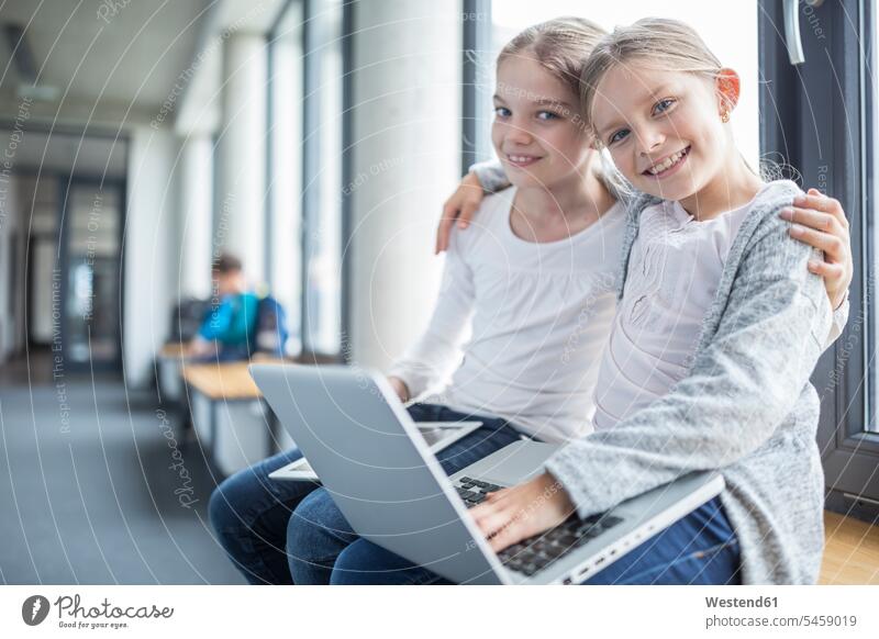 Portrait of two smiling schoolgirls with laptop and tablet embracing corridor hallway corridors hallways schools female pupils School Girl School Girls sitting