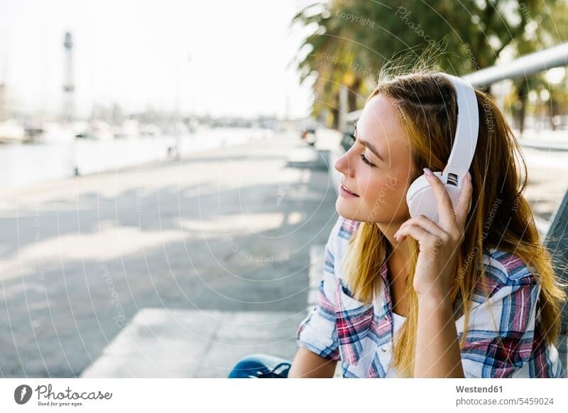 Young woman listening music through headphone sitting on footpath during sunny day color image colour image outdoors location shots outdoor shot outdoor shots