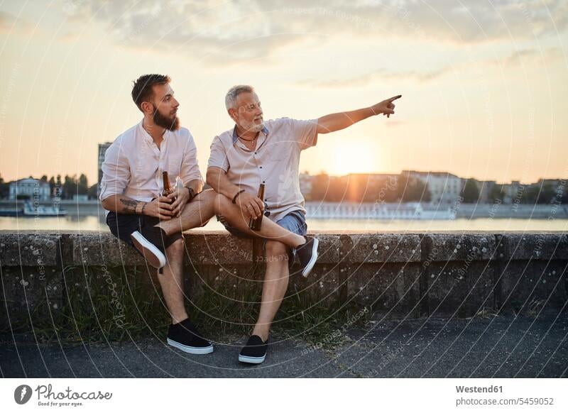 Father and adult son sitting on a wall at the riverside at sunset drinking a beer human human being human beings humans person persons caucasian appearance