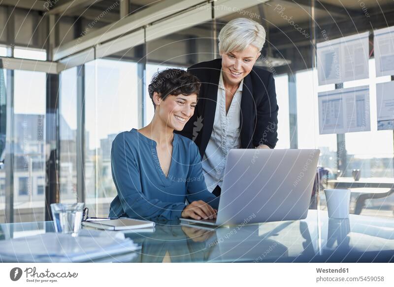 Two smiling businesswomen sharing laptop at desk in office desks smile offices office room office rooms Laptop Computers laptops notebook Female Colleague