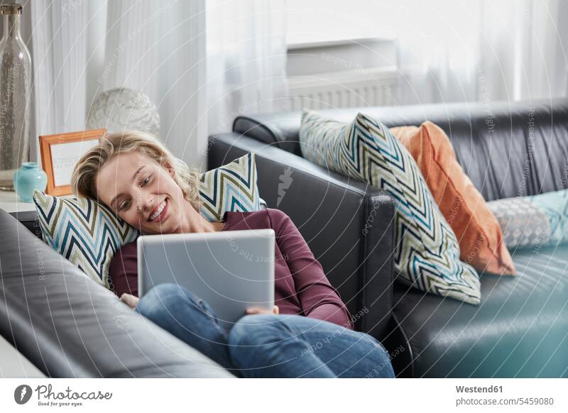 Smiling young woman with tablet lying on couch at home laying down lie lying down smiling smile females women settee sofa sofas couches settees digitizer