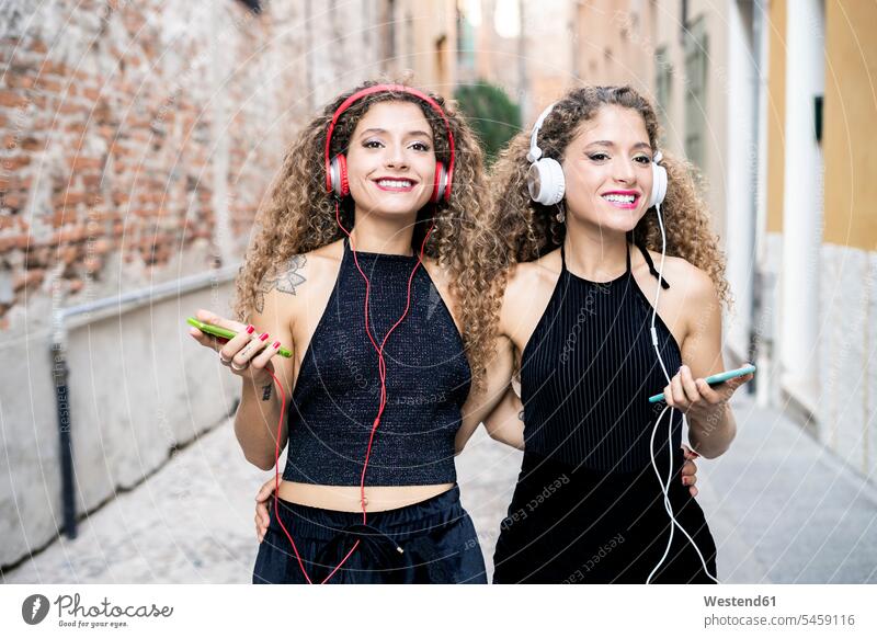 Portrait of smiling twin sisters listening music with headphones and cell phones in the city headset portrait portraits Smartphone iPhone Smartphones town